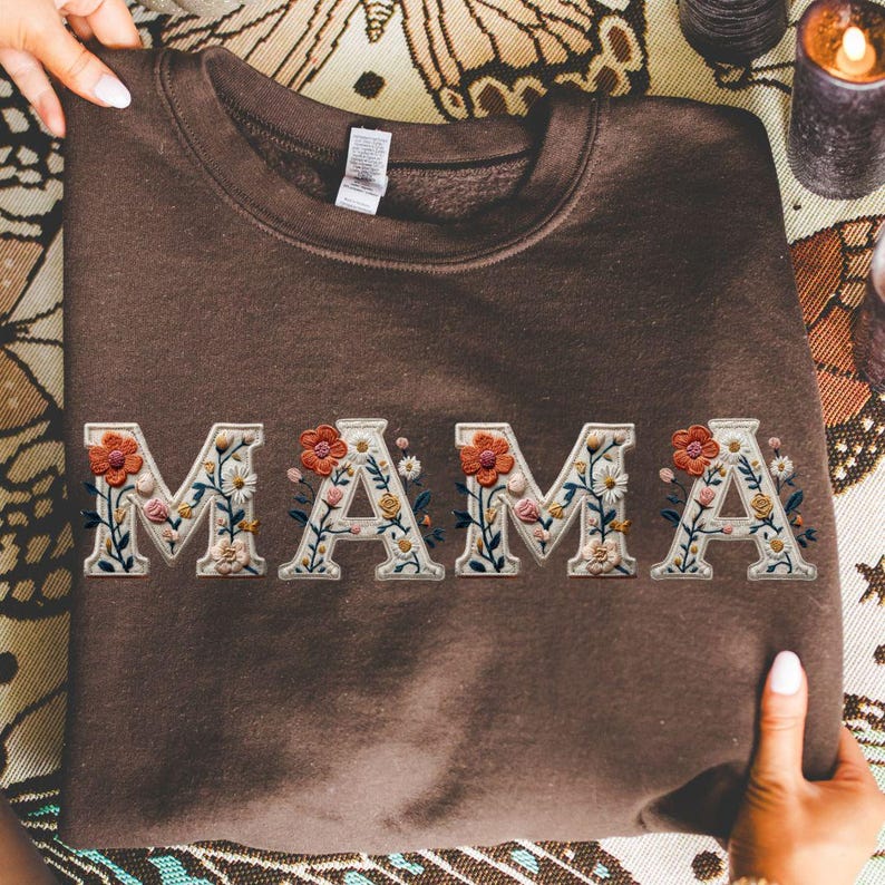 May include: A dark brown crewneck sweatshirt featuring the word MAMA embroidered in large, cream-colored block letters. Each letter is intricately filled with a colorful floral pattern consisting of orange, yellow, and blue flowers with green stems.