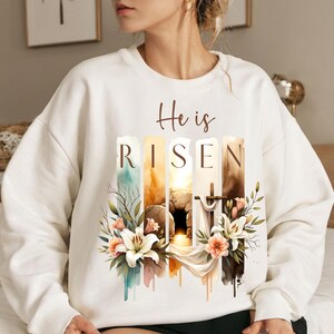 Puede incluir: Sudadera blanca con un dise&ntilde;o de acuarela de una tumba, una cruz y flores. El texto "He is Risen" est&aacute; impreso en la sudadera.