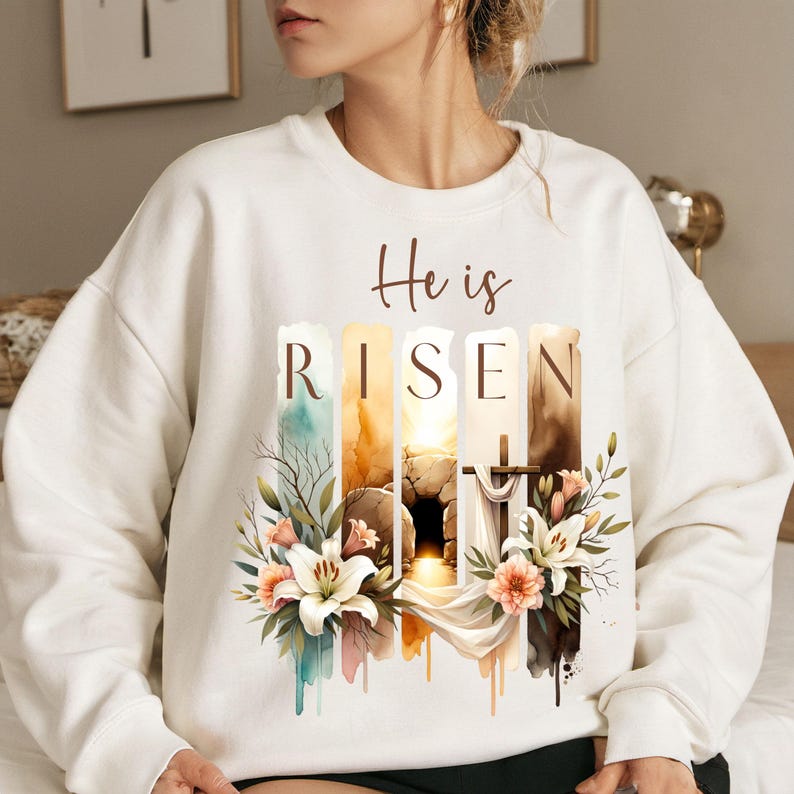 Puede incluir: Sudadera blanca con un dise&ntilde;o de acuarela de una tumba, una cruz y flores. El texto "He is Risen" est&aacute; impreso en la sudadera.