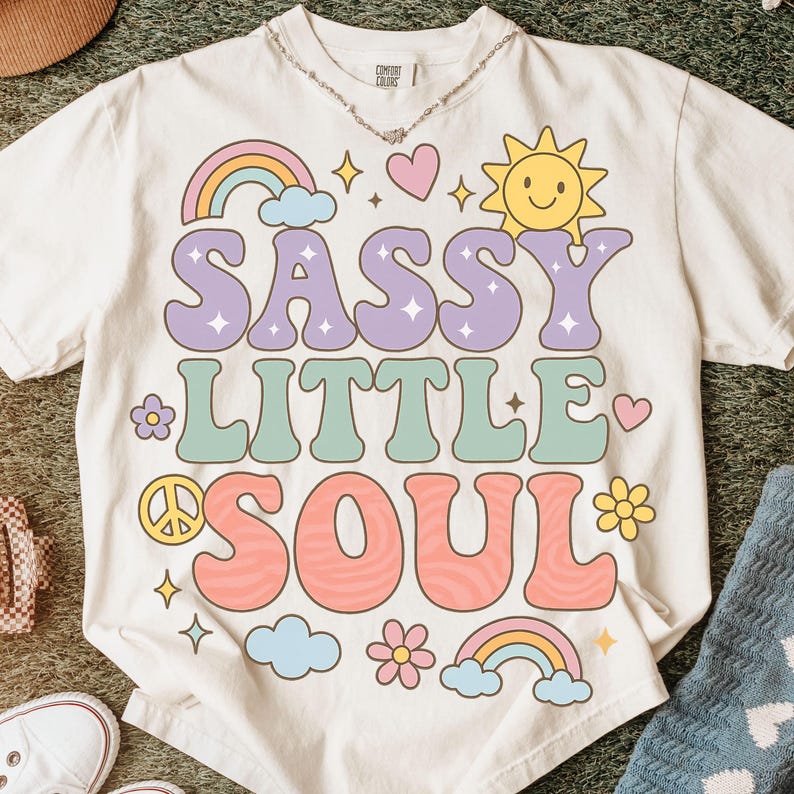 May include: An off-white short-sleeve t-shirt featuring the text SASSY LITTLE SOUL in large, wavy, multi-colored letters. The design includes a yellow sun, two rainbows, flowers, a peace sign, hearts, and small stars in pastel colors.
