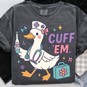 May include: Dark gray sweatshirt featuring a cartoon duck dressed as a nurse, holding a thermometer and stethoscope. The duck is wearing a nurse's cap and carrying a teal medical bag. The text "CUFF 'EM." is printed in purple and pink.