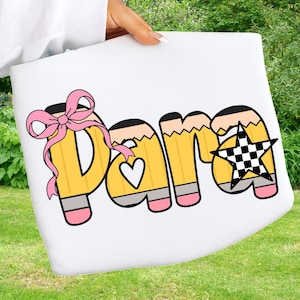 May include: White tote bag with the word "Para" in yellow pencil-shaped letters. The letter "D" has a pink bow, the letter "a" has a heart, and the letter "a" has a black and white checkered star.