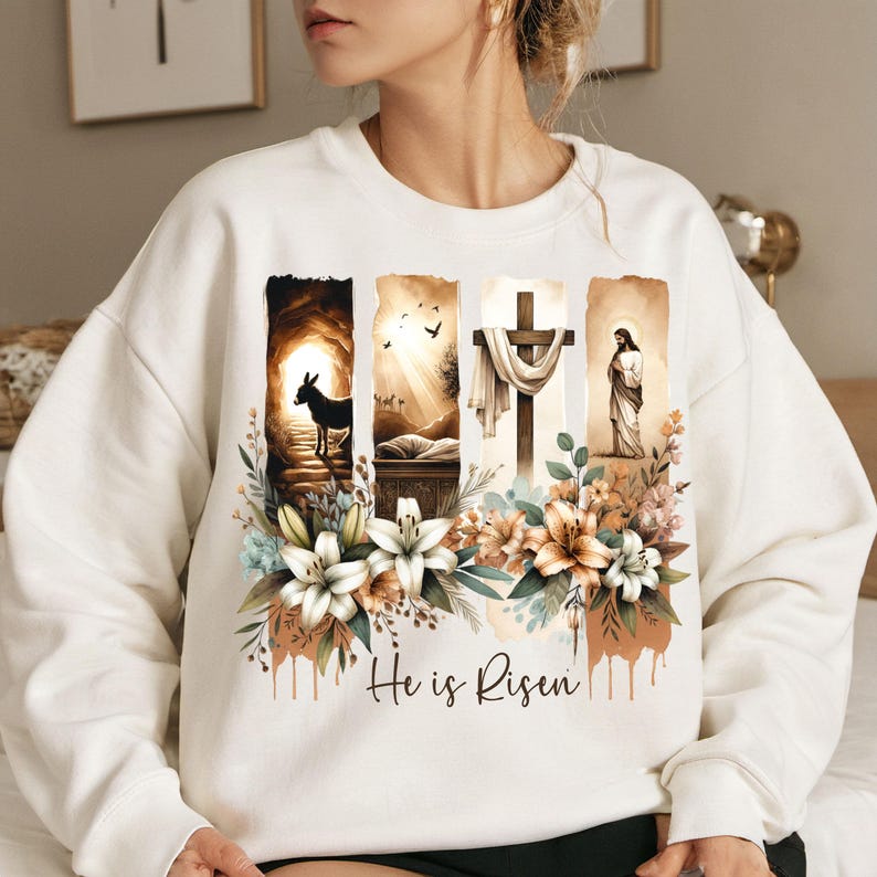 May include: White sweatshirt featuring a religious graphic with floral accents. The design includes images of a donkey, a tomb, a cross, and a figure, with the text "He is Risen" in a script font.