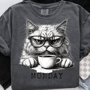 May include: Dark gray sweatshirt featuring a grumpy cat wearing glasses and holding a coffee cup. The cat illustration is in black and white. The word "MONDAY" is printed below the cat and cup.