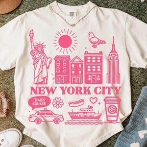 May include: A cream-colored short-sleeve t-shirt featuring a vibrant pink graphic print of New York City landmarks, including the Statue of Liberty, a pigeon, skyscrapers, a taxi, a ferry, and a coffee cup, with the text NEW YORK CITY.