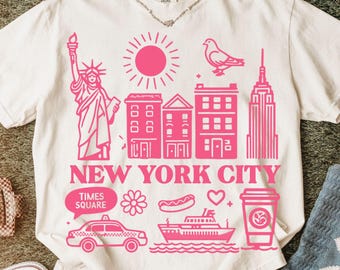 May include: A cream-coloured short-sleeve t-shirt featuring a vibrant pink graphic print of New York City landmarks, including the Statue of Liberty, a pigeon, skyscrapers, a taxi, a ferry, and a coffee cup, with the text NEW YORK CITY.