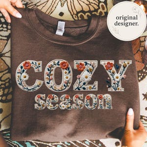 May include: A brown sweatshirt with the words "COZY SEASON" embroidered in a floral design. The letters are filled with white daisies, orange flowers, and green leaves. The sweatshirt is held up by a person.