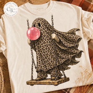 May include: A cream-colored t-shirt featuring a graphic of a ghost covered in a leopard print sheet, sitting on a wooden swing while blowing a large pink bubble. The ghost has two dark eye holes and is suspended by two metal chains.