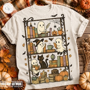 May include: A cream-coloured t-shirt featuring a graphic illustration of a dark metal bookshelf filled with books, pumpkins, a black cat, and three white ghosts. One ghost writes with a quill, another wears a witch hat, and the third reads a book.