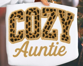 May include: A white sweatshirt featuring the word COZY in large, block-style letters with a brown and black leopard print pattern, followed by the word Auntie in a smaller, cursive-style font in a solid tan color embroidered on the fabric.