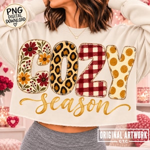 May include: A cream-colored cropped sweatshirt featuring the word COZY in large block letters with varied patterns: floral, leopard print, red and white buffalo plaid, and gold polka dots. The word season is written below in gold script.