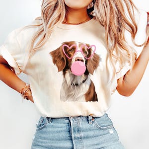 May include: A white t-shirt with a brown and white dog wearing pink heart-shaped sunglasses and blowing a pink bubble. The dog has brown eyes and a white patch on its chest.