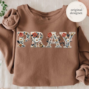 Puede incluir: Una sudadera marrón con la palabra "PRAY" bordada en letras blancas decoradas con flores coloridas. La sudadera tiene cuello redondo y mangas largas. Las palabras "original designer" están en un círculo en la parte superior derecha.