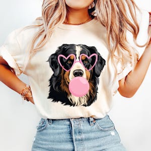 May include: Cream-colored t-shirt featuring a graphic of a Bernese Mountain Dog wearing pink heart-shaped glasses and blowing a pink bubble. The dog has black, brown, and white fur. The shirt is made of a soft material and is suitable for casual wear.