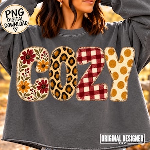 May include: A dark gray crewneck sweatshirt featuring the word COZY in large letters. The C has a floral pattern, the O has a leopard print, the Z has a red and white buffalo plaid pattern, and the Y has a yellow polka dot pattern.