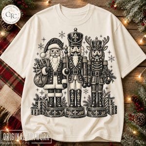 Op de afbeelding: Een crèmekleurig t-shirt met een zwart-witte grafische print van drie notenkrakerfiguren. De figuren omvatten een notenkraker in de vorm van de Kerstman, een traditionele soldaat en een rendier, omringd door diverse sneeuwvlokken.