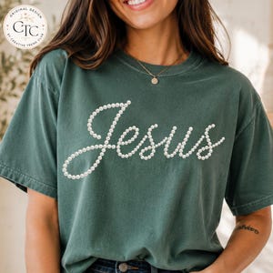May include: A dark green short-sleeve t-shirt featuring the word Jesus written in a cursive script made of white pearl-like beads. The shirt has a crew neckline and a relaxed fit, with a small circular logo in the top left corner reading CTC.