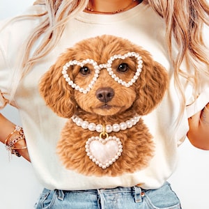 Peut inclure: T-shirt crème avec une illustration de caniche marron. Le caniche porte des lunettes en perles en forme de cœur et un collier de perles avec un pendentif en forme de cœur. La fourrure du chien est marron foncé, et le motif est centré sur le t-shirt.