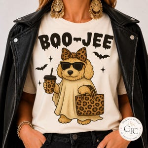 May include: A cream-colored t-shirt featuring a graphic of a golden dog wearing a ghost costume, sunglasses, and a leopard print bow. The dog holds a leopard print cup and tote bag. The text BOO-JEE is printed above in black, with small bat icons.