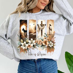 May include: A light gray sweatshirt featuring a religious graphic with four vertical panels. The panels depict scenes with a donkey, a tomb, a cross, and a figure. Floral accents and the text "He is Risen" complete the design.