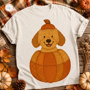 以下が含まれることがあります： クリーム色の半袖Tシャツ。小さなカボチャの帽子をかぶり、様々な質感の模様が施された大きなオレンジ色のセグメント化されたカボチャの中に座っている、巻き毛のゴールデンブラウンの犬のグラフィックが中央にプリントされています。