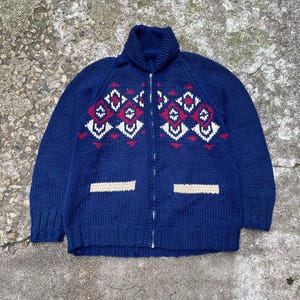 May include: A blue knitted cardigan with a zipper closure, featuring a geometric pattern in red, white, and cream on the front. The cardigan has two patch pockets with white trim.
