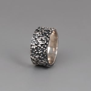 May include: A textured silver ring with a rough, uneven surface. The ring has a wide band and a smooth, polished interior. The ring is set against a gray background.