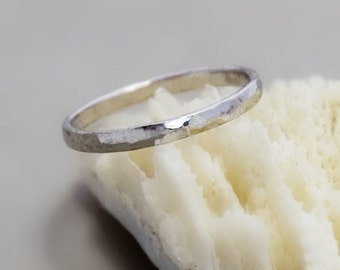 May include: A thin silver band ring with a hammered texture. The ring is sitting on a white coral-like surface.