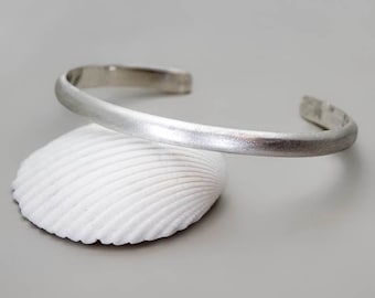 May include: A silver bangle bracelet with a brushed finish. The bracelet is open and has a simple, minimalist design.
