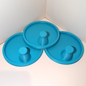 May include: Three circular, bright blue plastic air hockey pushers. Each pusher features a flat, round base with a textured, perforated surface and a central, raised handle with a ridged, scalloped top edge for a secure grip.