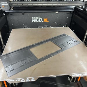 May include: A black 3D-printed part with a rectangular frame and internal cutouts, resting on a brown print bed. The printer is an Original Prusa XL, with multiple print heads visible above.