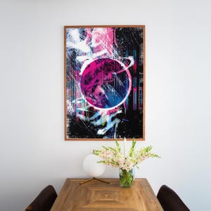 May include: Abstract art print in a wooden frame. The artwork features a vibrant design with a large pink circle and a black background with splashes of blue, white, and pink. The print is displayed on a white wall above a wooden table.