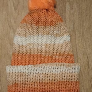 May include: A knitted beanie in shades of orange, cream, and white, topped with a fluffy orange pom-pom. The hat features a folded brim and a striped pattern. This warm winter hat is perfect for cold weather.