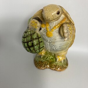 May include: A ceramic turtle figurine with a tan shell and a gold necklace. The turtle is holding a green bag with a grid pattern. The figurine is standing on a base with green and brown details.