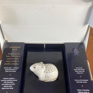 Könnte beinhalten: Keramik-Mausfigur der Royal Crown Derby Collectors Guild. Die Maus ist weiß mit braunen und goldenen Akzenten, präsentiert in einer marineblauen Box. Die Box enthält Text über die Vorteile der Mitgliedschaft in der Gilde.