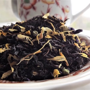May include: A close-up of a pile of loose leaf black tea with dried fruit and flower petals. The tea is in a white ceramic bowl with a brown floral pattern.