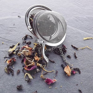 May include: A silver tea infuser with a chain and a loose leaf tea blend of black tea, rose petals, and dried fruit pieces scattered on a gray surface.