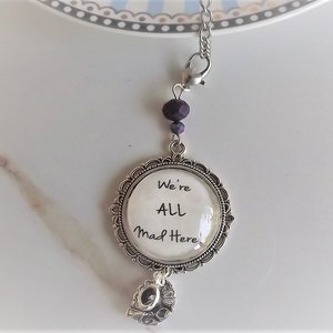 May include: A silver charm necklace with a round pendant featuring the text "We're All Mad Here". The pendant is attached to a silver chain with a purple bead.