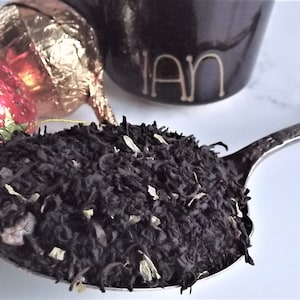 May include: A silver spoon filled with loose leaf black tea with white flower petals. The tea is next to two wrapped chocolate candies, one red and one gold.