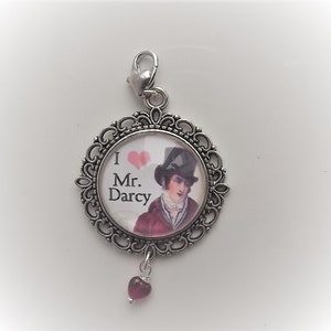 May include: Silver charm with a round glass cameo featuring a portrait of Mr. Darcy from Pride and Prejudice. The cameo is surrounded by an ornate silver frame and has a small red heart charm hanging from the bottom.