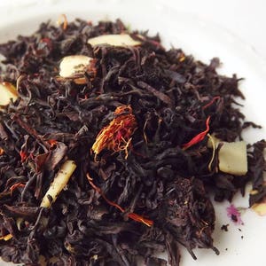 May include: A close-up of a blend of black tea leaves, dried fruit pieces, and almond slivers. The tea blend is a mix of dark brown, red, and beige colors.