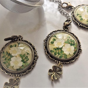 May include: Three gold-toned tea infuser charms with white flowers and green leaves inside glass domes. Each charm has a four-leaf clover dangling below.