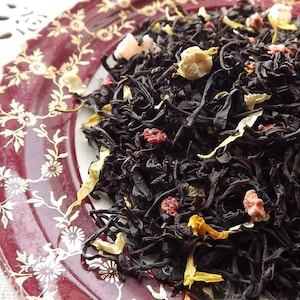 May include: A close-up of black tea leaves with dried fruit pieces, including yellow citrus peel and pink fruit pieces. The tea is on a red and gold patterned plate.