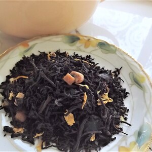 May include: A close-up of a white ceramic plate with a pile of black tea leaves, orange peel, and small orange-colored pieces. The tea leaves are loose and appear to be a blend of black tea and fruit.