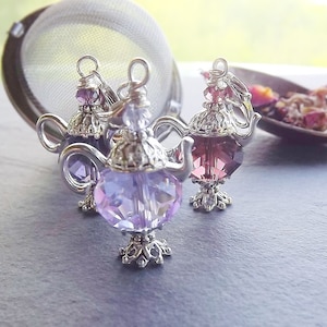 May include: Three decorative tea infusers shaped like teapots, each with a different shade of purple crystal. The infusers have silver-colored metal accents and are placed near a stainless steel tea strainer. A spoon with loose tea is in the background.