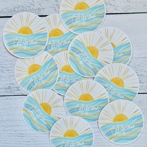 May include: A set of 10 round stickers featuring a stylized sun rising over blue and yellow waves with the word "California" written in white script.