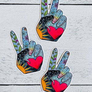May include: Three peace sign stickers with a hand-drawn design. Each sticker features a red heart, a black and gold sunburst, and a blue background with flowers and leaves. The stickers are on a white wooden surface.