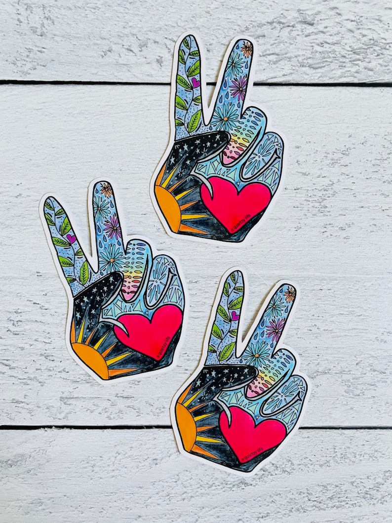 May include: Three peace sign stickers with a hand-drawn design. Each sticker features a red heart, a black and gold sunburst, and a blue background with flowers and leaves. The stickers are on a white wooden surface.