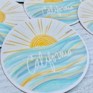 May include: A round sticker with a blue and gold watercolor design. The sticker features a stylized sun with rays and the word "California" written in white script. The text "A Charity Life" is printed in small font at the bottom of the sticker.