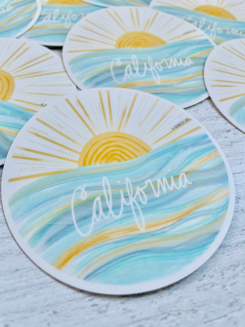 May include: A round sticker with a blue and gold watercolor design. The sticker features a stylized sun with rays and the word "California" written in white script. The text "A Charity Life" is printed in small font at the bottom of the sticker.